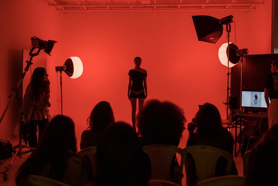 A fashion model poses in a studio with dramatic red lighting while an audience observes the scene.